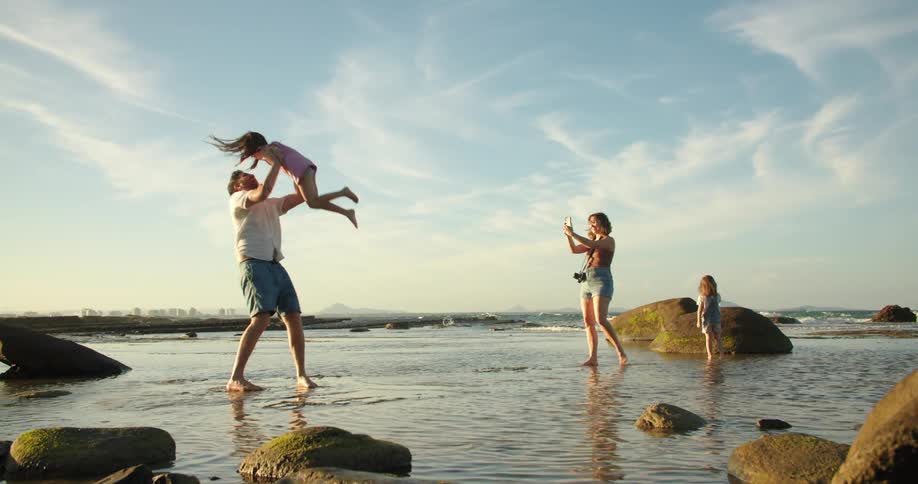 Mother takes video with smartphone as one of her daughters runs across beach to be picked up by her father