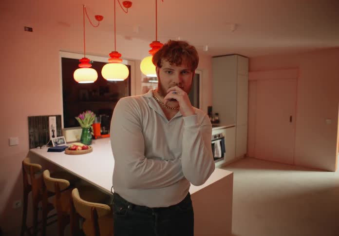A man is standing in a room near the counter and chairs with his hand under his chin