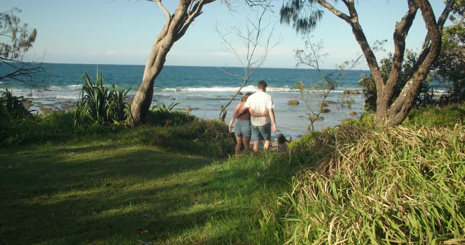 Husband and wife are walking across the grass with their arms around each other as their two young daughters run down the hill towards the beach