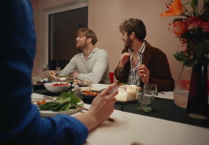 A small group of people are sitting down eating and talking and there is a bouquet of flowers on the table