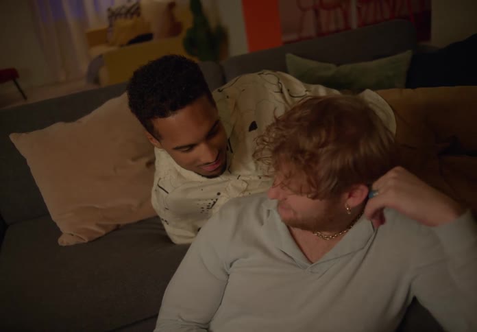 A man plops down on the couch behind a man sitting on the floor