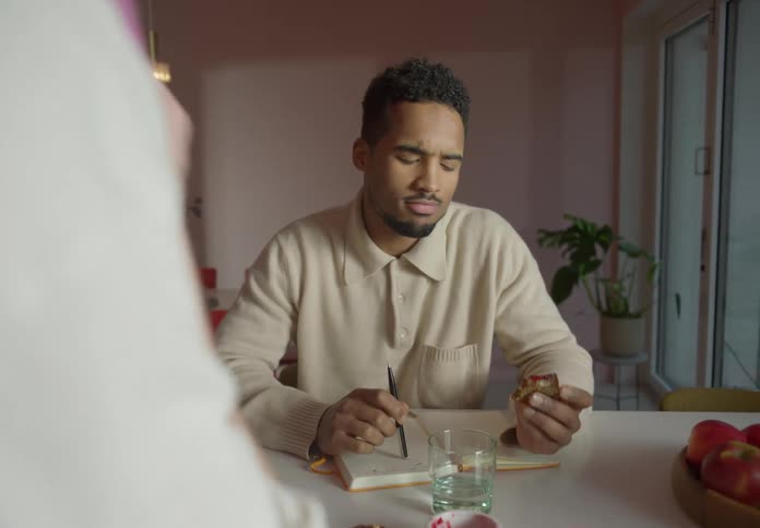 A man eating and writing while someone talks off screen