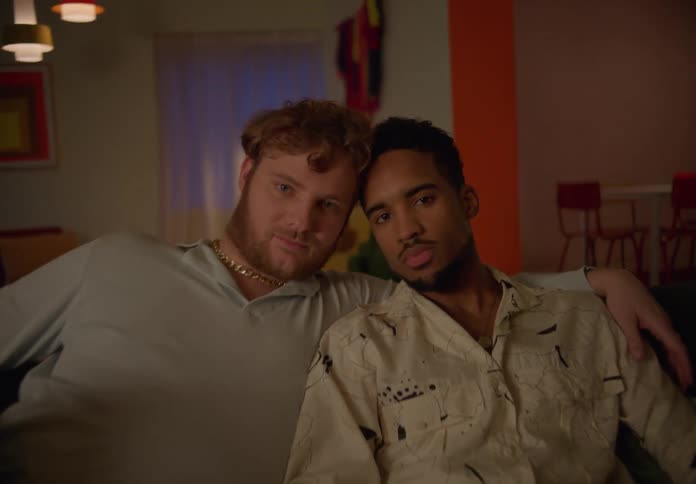 Gay couple sitting on couch looks at camera and holds each other in living room