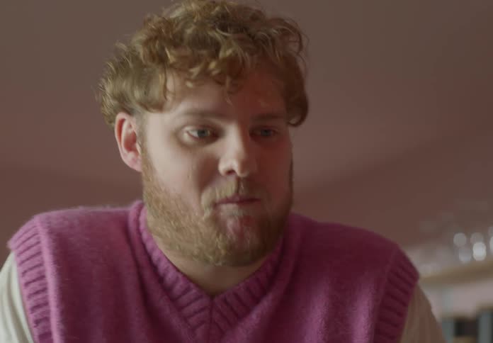 A man takes a bite of a piece of toast with jam on it while standing indoors during the day