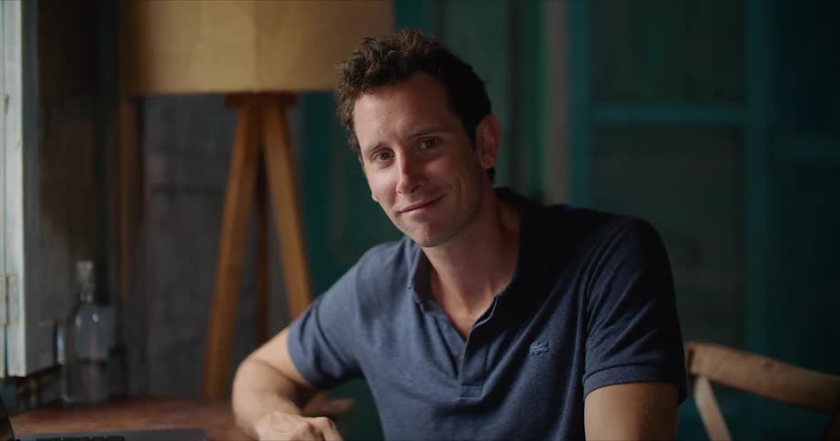 A man is sitting at a desk with an open book and a laptop computer as he looks up and smiles