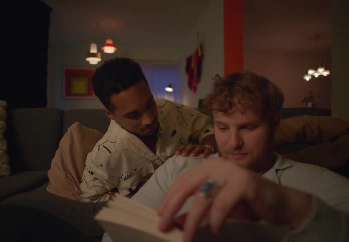 Gay man reading book is being stroked by his partner sitting on couch in the living room