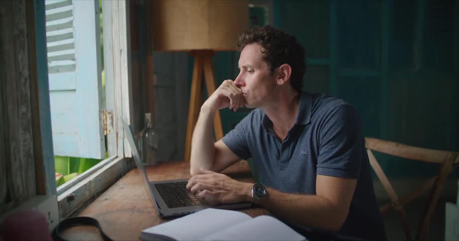 A man is sitting at a desk in front of a window looking at an open book as he types on a laptop computer