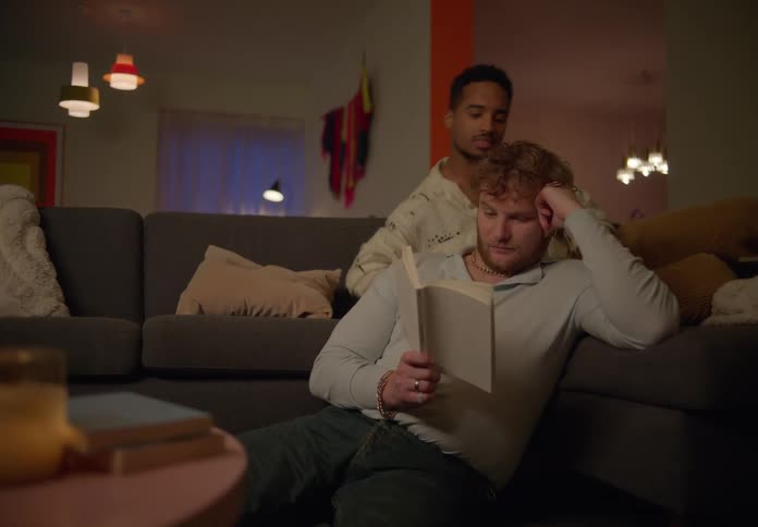 A man gets on the couch beyond his boyfriend reading a book