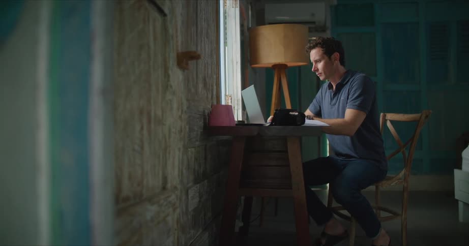 A man is sitting at a desk in front of a window turning pages in an open book as he types on a laptop computer
