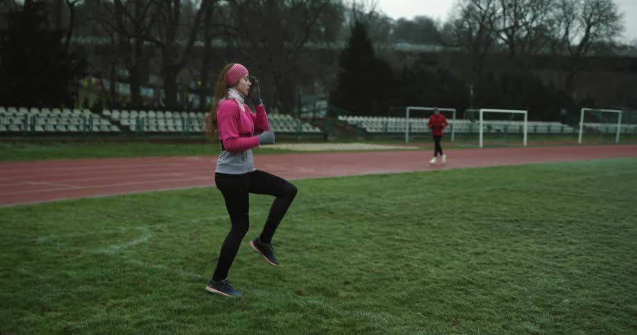 A woman is standing on the grass running in place as another person runs around the track
