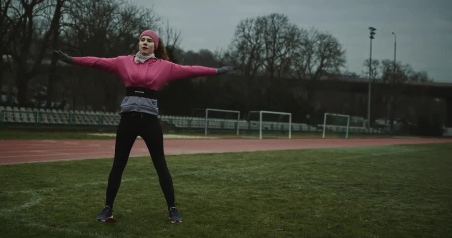 A woman is standing on a grass track field doing stretching exercises