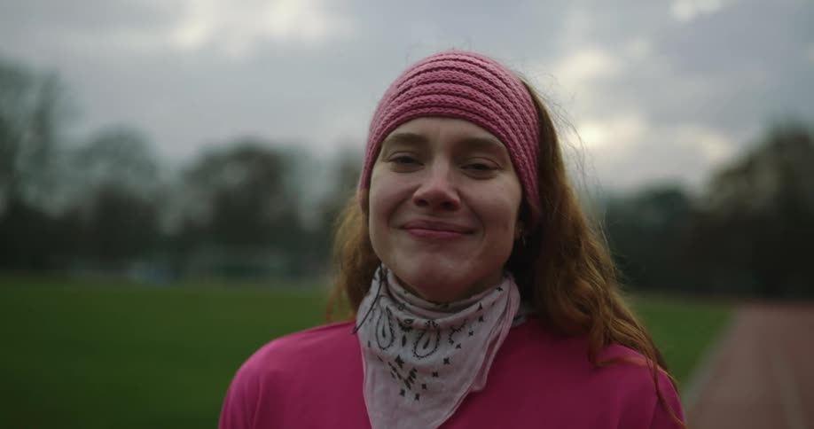 Woman with brown hair pink headband and pink top smiles at camera on running track