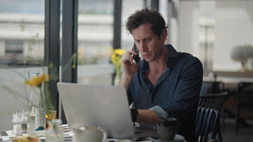 Man talks on smartphone while sitting with laptop at cafe
