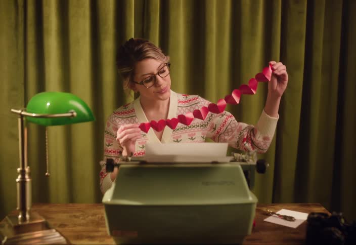 A woman sitting at a desk holds up a string of paper cutout hearts while indoors