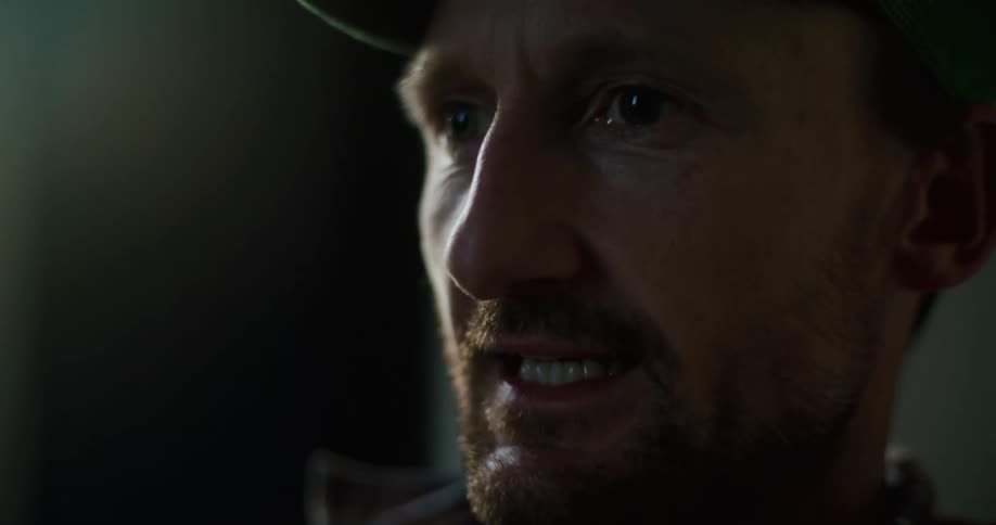 Cu man with red beard and baseball cap having a dialog with somebody off camera explaining and then pauses to smoke a pipe or vape