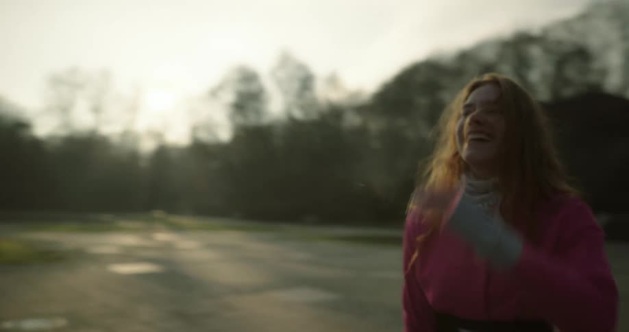 A teenage girl is standing outside in a field with trees in the distance as she breathes deeply and her breath is seen in the cold