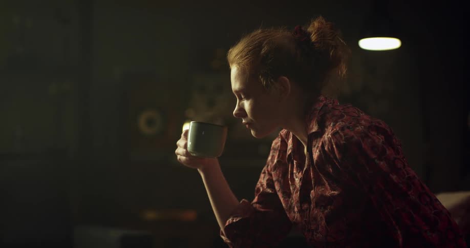 Redheaded woman sits up to take sip from steaming mug before sitting back while looking up on couch
