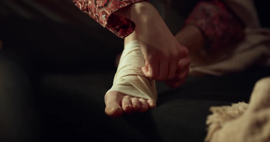 A woman is wrapping her foot with a bandage while sitting indoors during the day