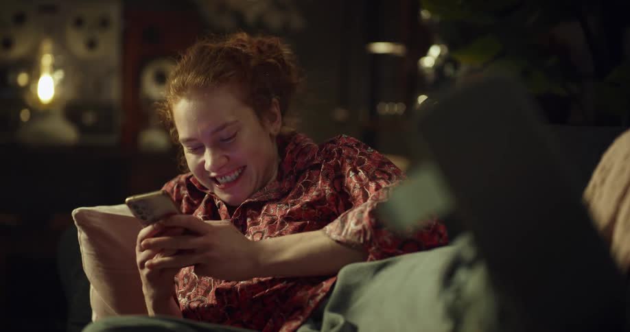 Young woman with red hair lying on couch texting and laughing elevating an injured legfoot