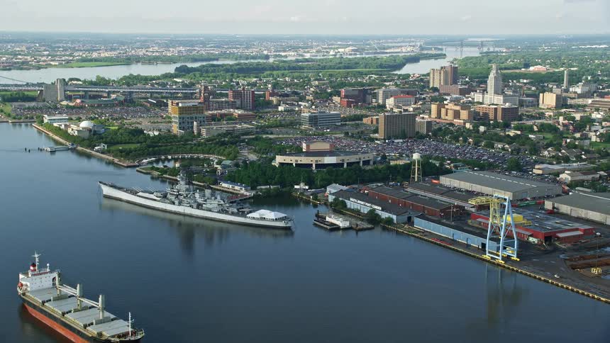 An aerial view of the USS New Jersey and other naval and industrial ships approaching Camden New Jersey with partly cloudy skies and a crowd gathered at the BB&T Pavilion