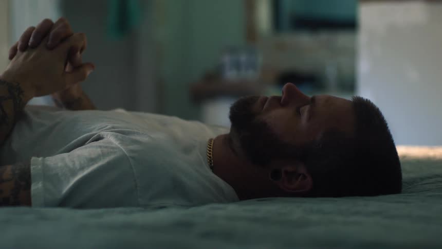 Man with beard tattoo necklace and white t shirt gets out of bed praying