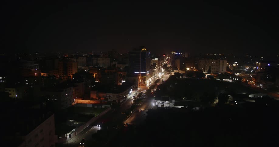 The city is lit up at night as the headlights of the cars light up the main road and lights in the windows of buildings light up all around