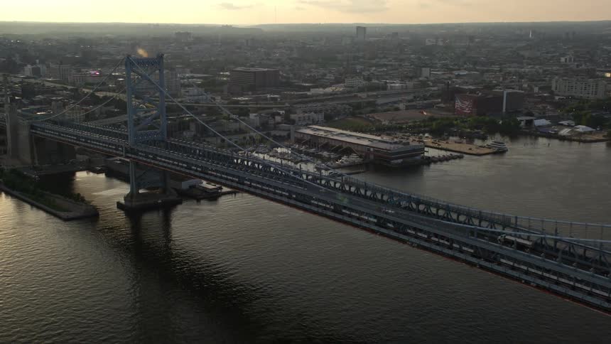 Approaching Benjamin Franklin Bridge at dusk spanning the Delaware River during sunset shot from an aerial 