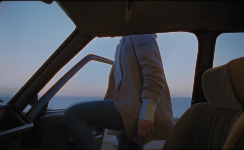 A woman enters the passenger seat of a vehicle parked on the coast in the evening