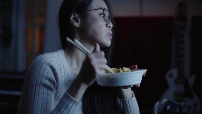 Woman eats noodles and works on computer reflected by her eyeglasses in a dim room