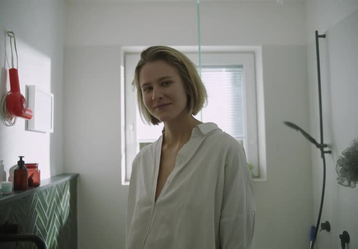 A woman with blond hair wearing a white shirt is standing in front of the bathroom mirror as she turns and smiles