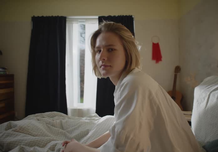 A woman sit on a messy bed near a bedroom window