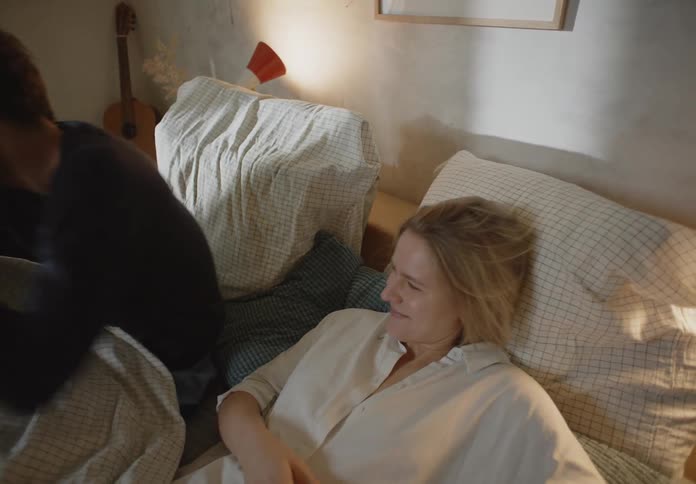 A woman on her phone while a man in the bed next to her kisses her on the forehead