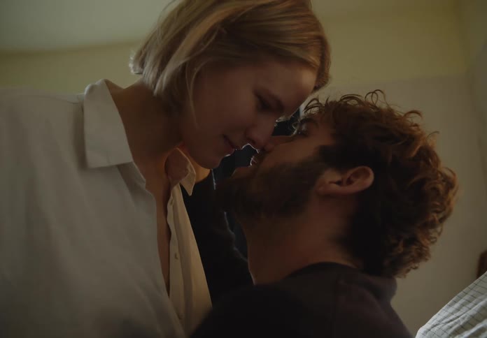 A woman stands leaning down looking at the man sitting in front of her as he reaches his hand up a brushes her hair back behind her ear than she leans in and they look at each other before embracing in a passionate kiss