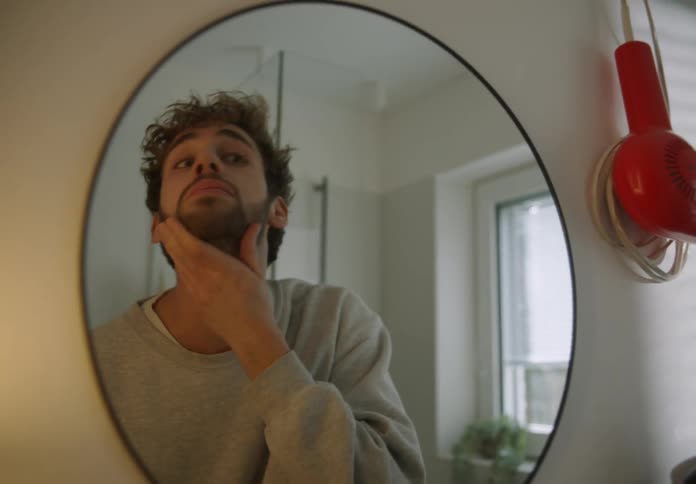 A man is inspecting his growing beard in a bathroom mirror at home in the morning