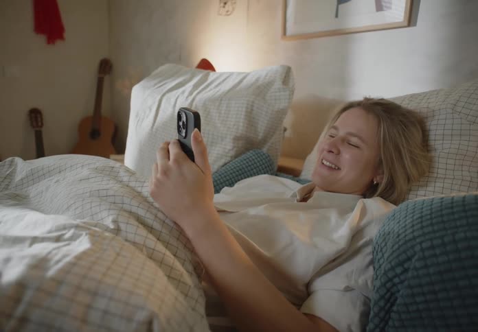 A young woman is laying in bed and laughing at something on her phone