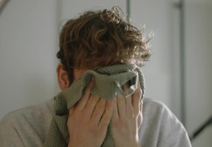 A man is standing in front of a bathroom mirror and drying his face in the daytime