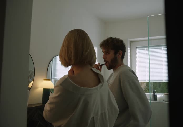 Man with beard and sweater and blonde woman with white shirt brush teeth in the bathroom