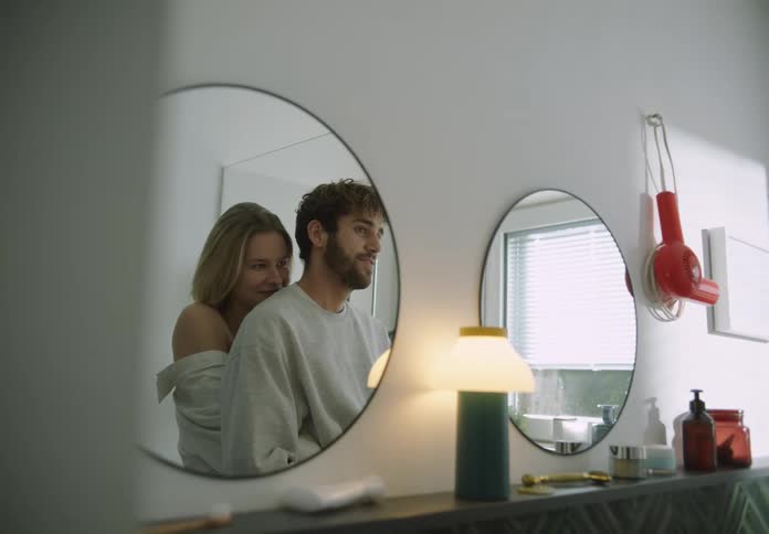 A man and woman embrace while standing in a bathroom and looking in the mirror
