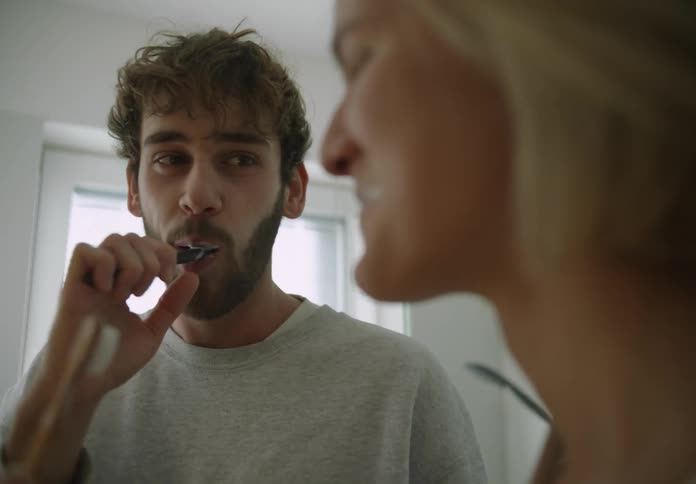 A man and woman brush their teeth in the bathroom while joking around