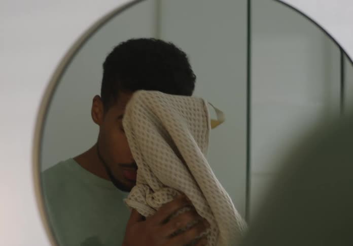 Man with beard and light blue t shirt dries his face with towel in bathroom