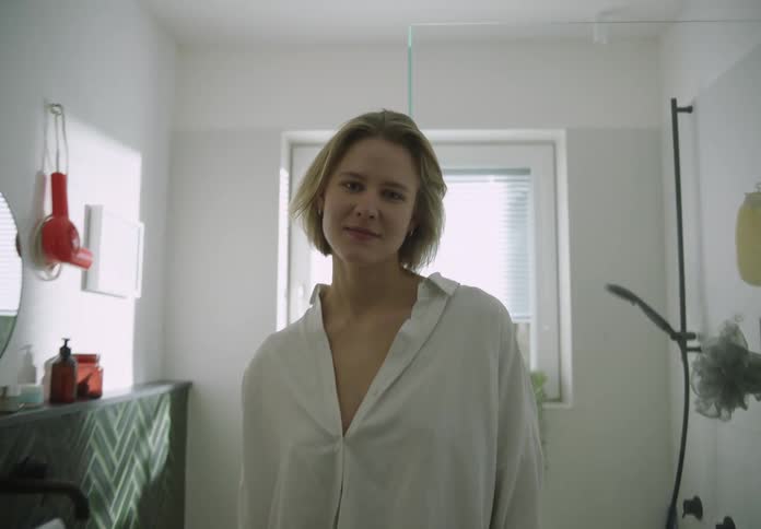 A woman with blond hair wearing a white shirt is standing in the bathroom as she steps forward and smiles