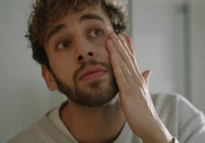 A man with a beard applies cream to his face using his hands while looking in the mirror