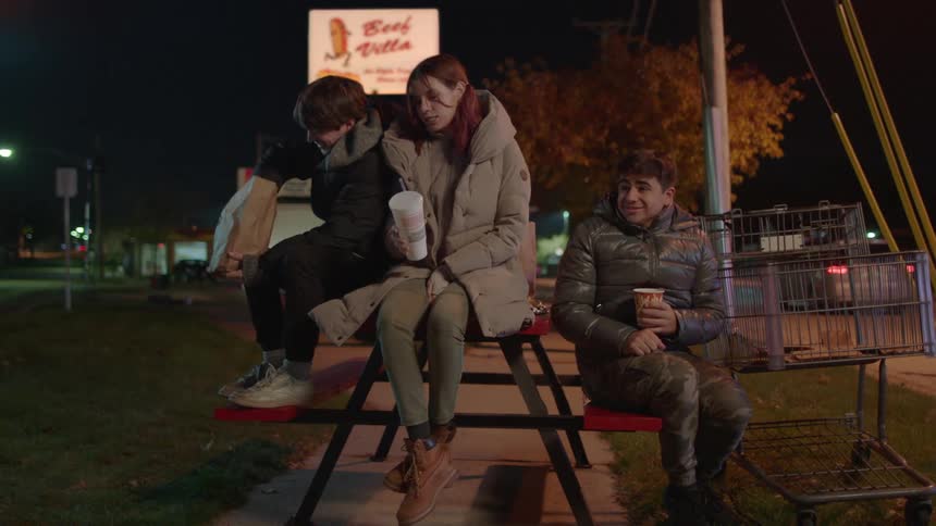 A teen girl and two boys sitting on a park bench at night laughing and enjoying some fast food and soda
