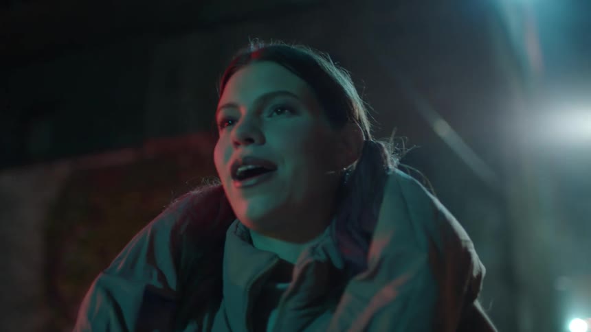 A woman wearing a winter coat is talking as she walks on a street at night and the street light shines brightly in the darkness