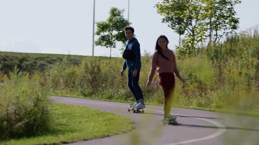 Two teenagers are riding their skateboards down a road on a hill surrounded by plants and weeds