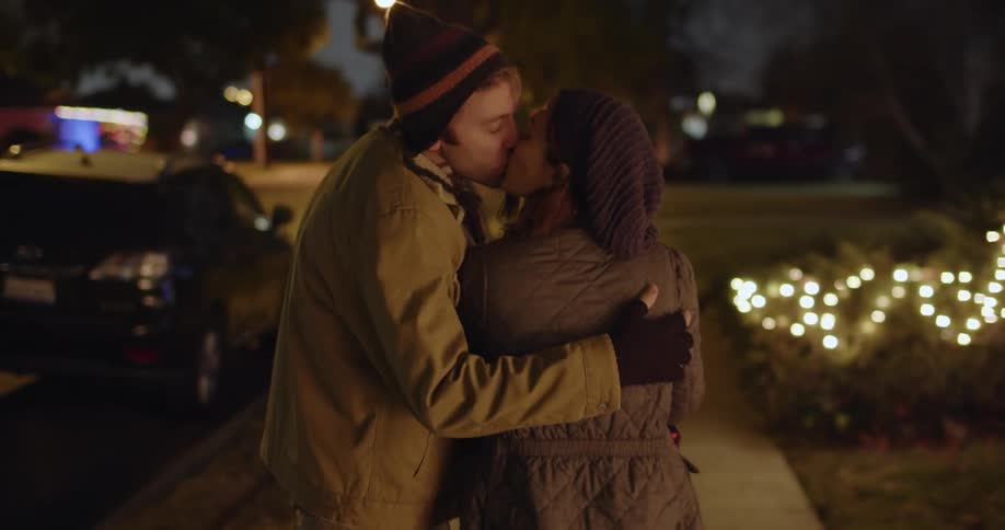 A man and a woman are walking on a neighborhood street looking at christmas lights and she is carrying wrapped presents as he puts his arm around her and they kiss