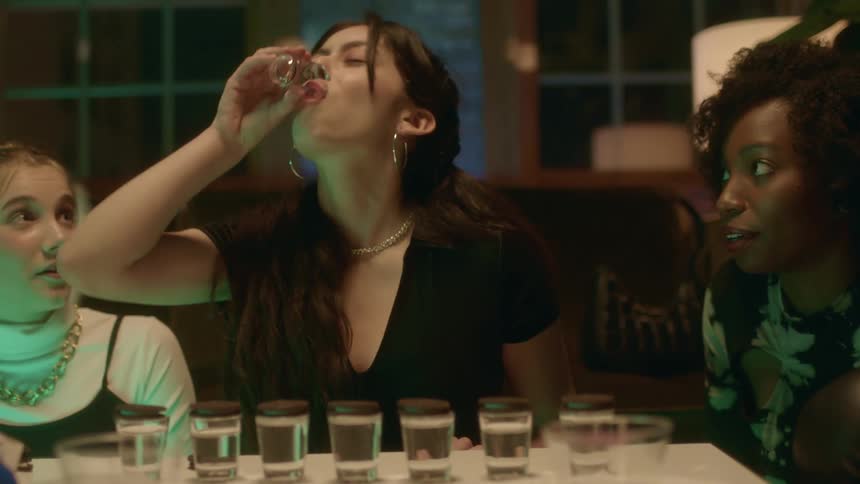 A young womans friends appear shocked as she downs multiple shots of alcohol