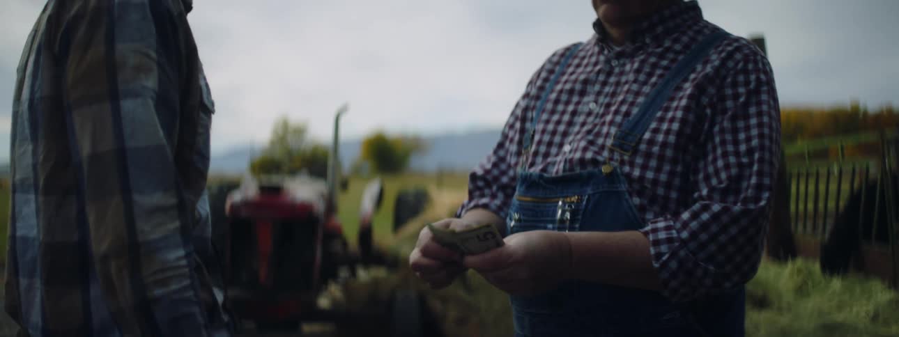 Farmer standing with tractor in background pays cash to man then shakes his hand