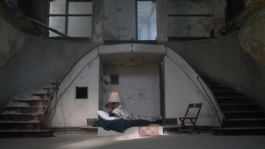 Person lies on bed between two flights of stairs in damaged building