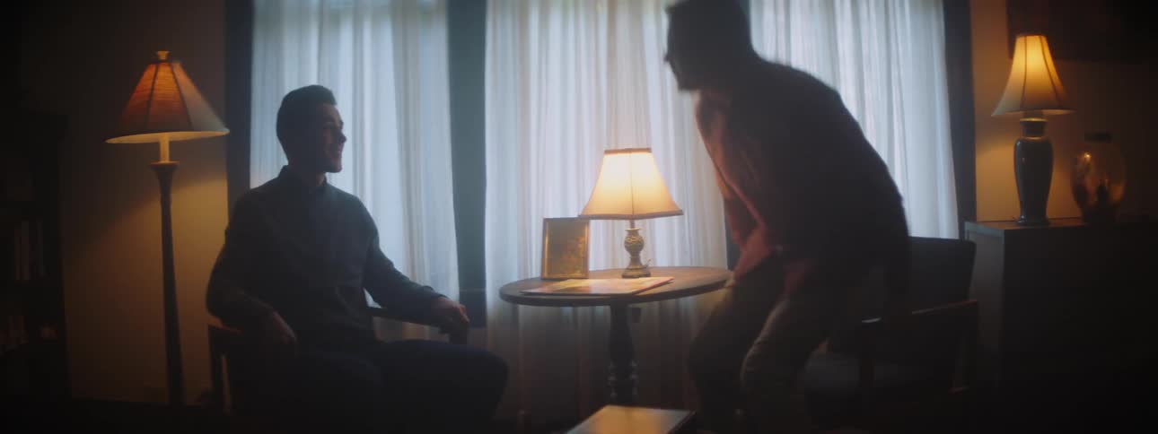 Two men sitting in a living room stand and shake hands and then embrace in the daytime